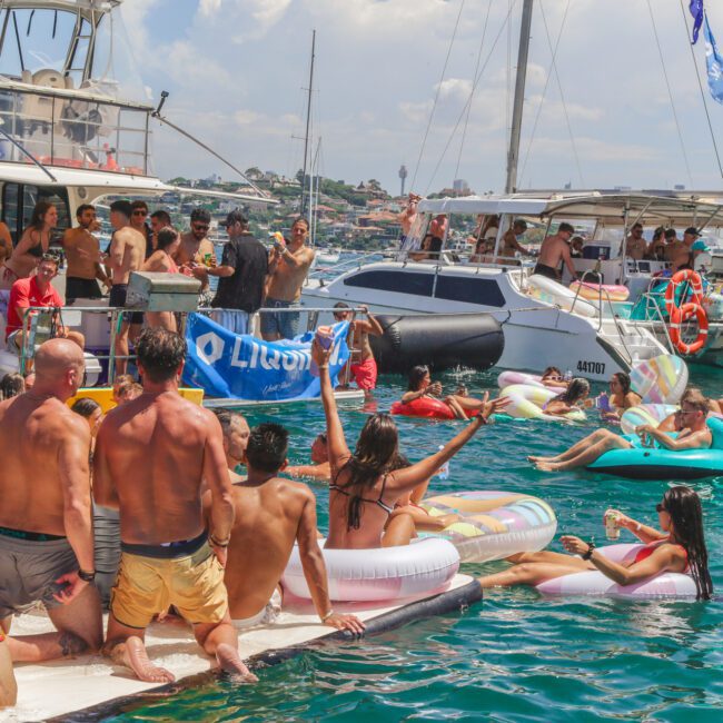 A lively group of people enjoy a boat party in the sun, with many on inflatables and floating platforms in clear blue water, surrounded by yachts and catamarans. Everyone appears to be celebrating and having fun.