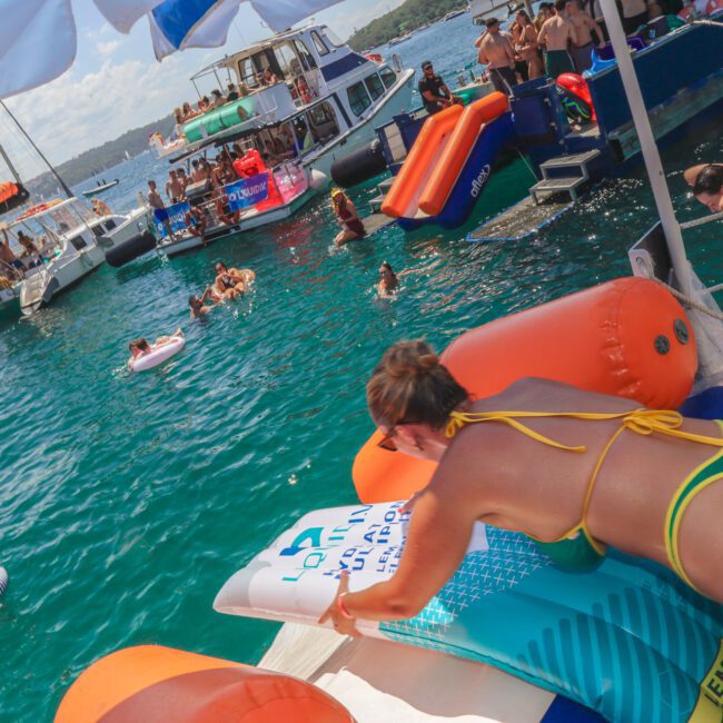 A woman in a green and yellow bikini climbs onto an inflatable float at a lively boat party. Several people relax on floats and swim in the blue water, while others gather on boats anchored nearby under a sunny sky.