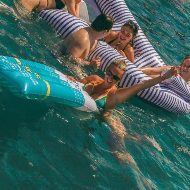 A group of people relax and laugh together on large inflatable floats in the ocean on a sunny day. The water is clear blue, and everyone appears to be enjoying the fun and social atmosphere.