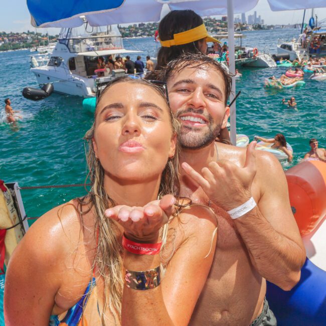 Two people smile at the camera on a boat, with the woman blowing a kiss and the man making a shaka hand sign. The background shows a lively water party with boats and people swimming and relaxing.