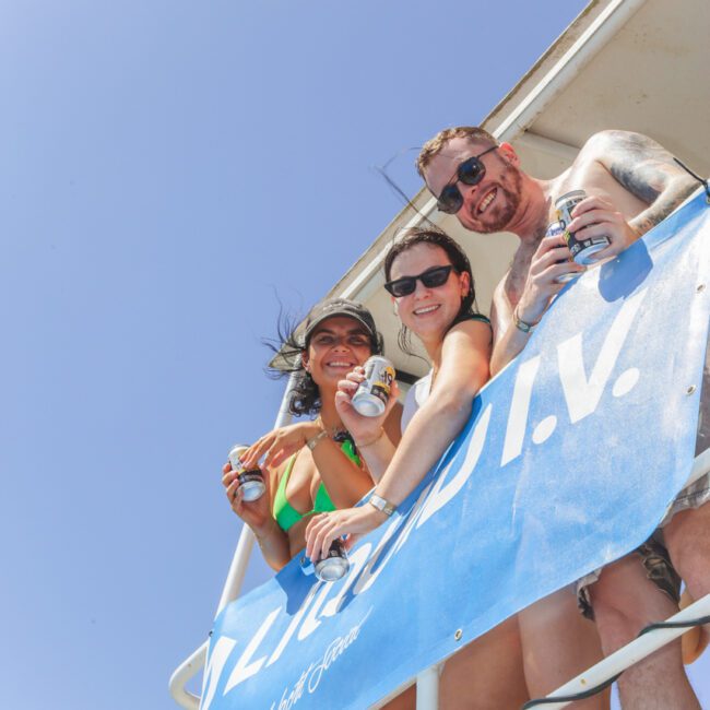 Three people in swimsuits smile and hold drinks while standing on a boat deck, looking down at the camera. A blue banner with white text is displayed on the railing. The sky is clear and sunny.