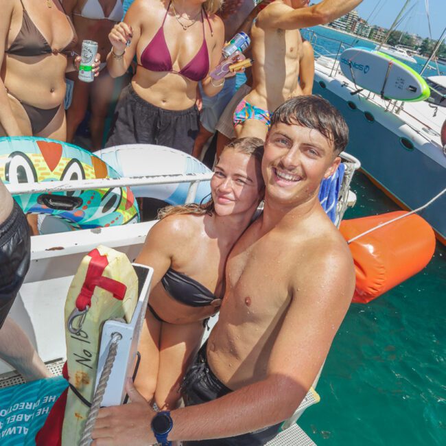 A group of people in swimsuits enjoy a sunny day on a boat, smiling and holding drinks. Two people stand at the boat’s ladder, posing for the photo with others socializing in the background on the deck.