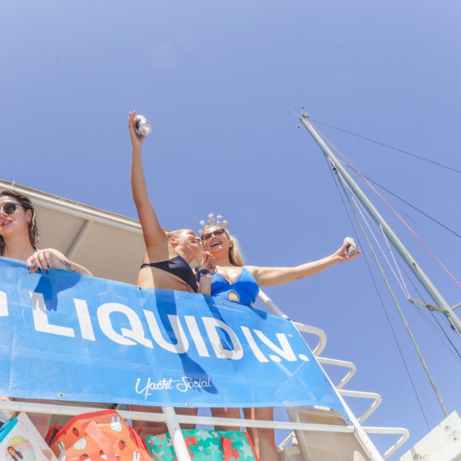 Four women in swimsuits stand on a boat deck, smiling and raising drinks, with a "LIQUID I.V." banner in front. The sky is clear and blue, and a sailboat mast is visible in the background.