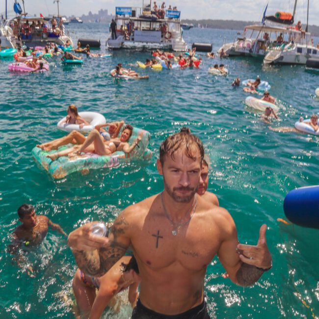 A shirtless man with tattoos stands in the water making a hand gesture at a crowded boat party, with people swimming, relaxing on inflatables, and yachts anchored in the background.