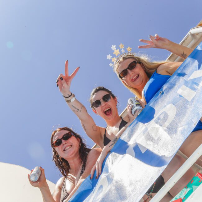 Three smiling women in swimsuits and sunglasses pose on a boat, one wearing a star crown and flashing a peace sign. They're enjoying a sunny day under a clear blue sky, standing behind a blue and white banner.