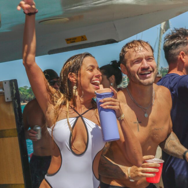A group of young adults in swimsuits laugh and cheer while holding drinks on a boat, enjoying a sunny day on the water. The atmosphere is lively and festive.