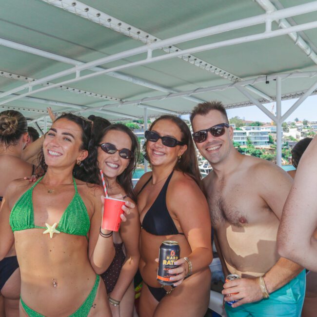 A group of four smiling people in swimsuits hold drinks and pose together on a boat during a sunny day, with other people and buildings in the background.