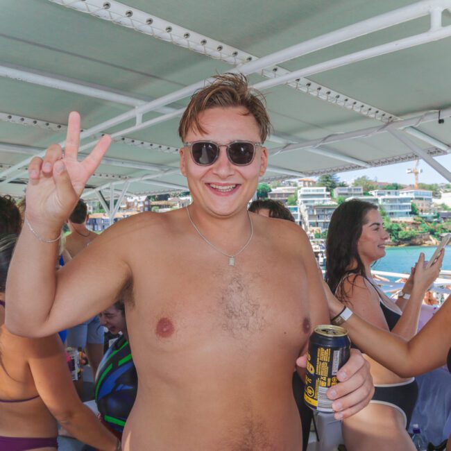 A shirtless man wearing sunglasses smiles and flashes a peace sign while holding a can of drink on a boat crowded with people in swimwear, with water and buildings visible in the background.