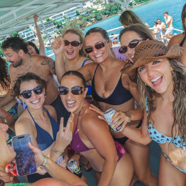 A group of smiling women in swimsuits pose together on a boat, enjoying a sunny day. Some hold drinks, one flashes a peace sign, and another takes a selfie. Blue water and coastal buildings are visible in the background.