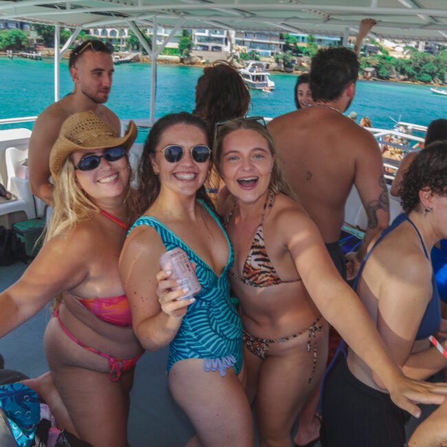 A group of people in swimsuits smile and pose together on a boat with turquoise water and a coastal town in the background, enjoying a sunny day.