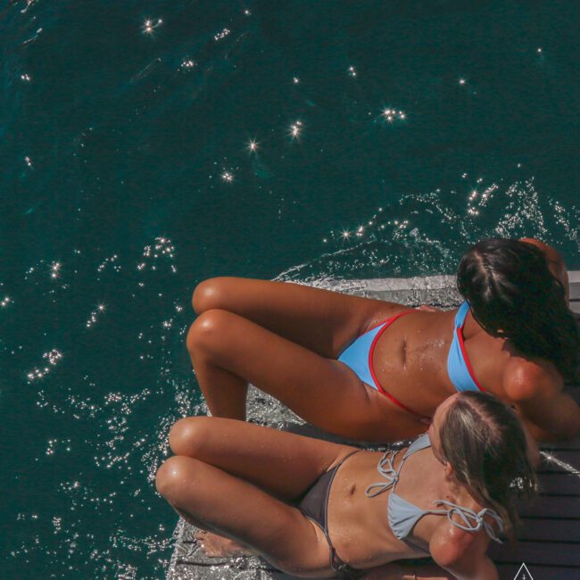 Two women in bikinis relax on a wooden platform by deep blue water, with sunlight reflecting off the surface. The women are sunbathing, partially wet, and the scene feels summery and leisure-filled.