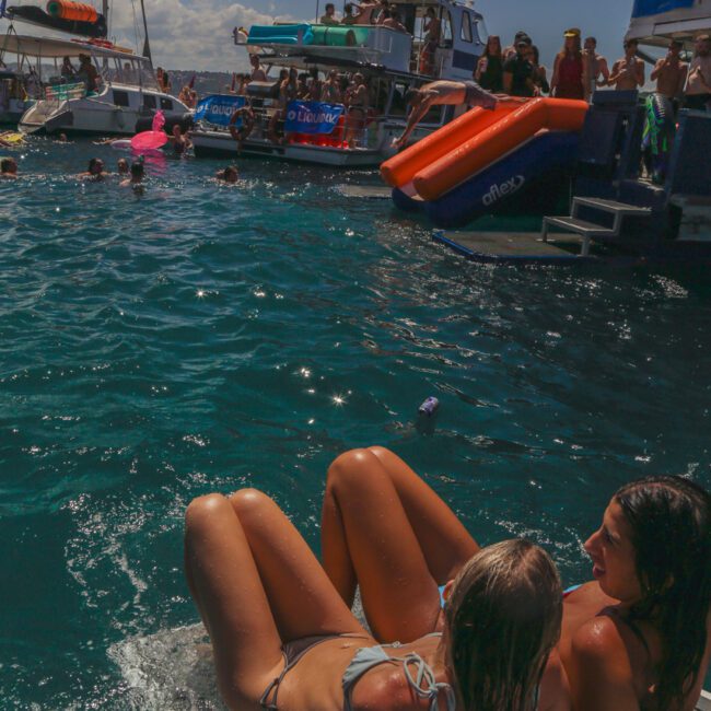 Two women in swimsuits relax on the edge of a docked boat in clear blue water, while several boats with people enjoying the sun and water can be seen in the background at a lively party scene.