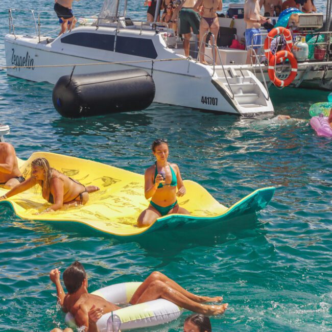 People enjoy a lively boat party on the water, some lounging on colorful floaties and mats, while others relax on nearby boats under the sun. The scene is festive with bright swimwear and vibrant inflatables.
