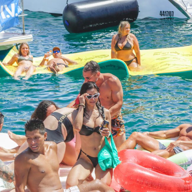 A group of people in swimsuits relax on colorful inflatables and floaties in clear blue water under bright sunlight, enjoying a lively summer gathering.