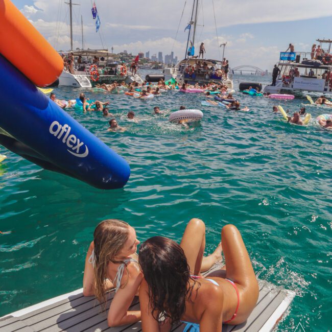 Two women in swimsuits relax on a dock in front of a crowded, lively harbor with boats, people swimming and lounging on inflatables, and a city skyline in the background under a partly cloudy sky.