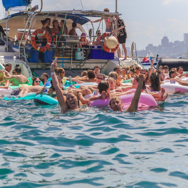 A large group of people on colorful inflatable floats relaxes and has fun in the water near boats, with a city skyline visible in the background under a bright sky.