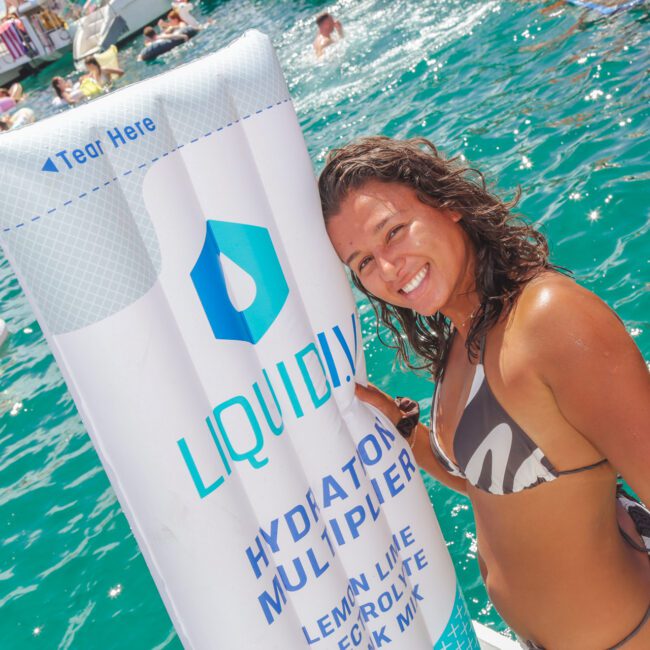 A woman in a swimsuit smiles while leaning against a large inflatable Liquid I.V. Hydration Multiplier packet in a crowded, sunlit swimming area with people floating in the water and boats nearby.