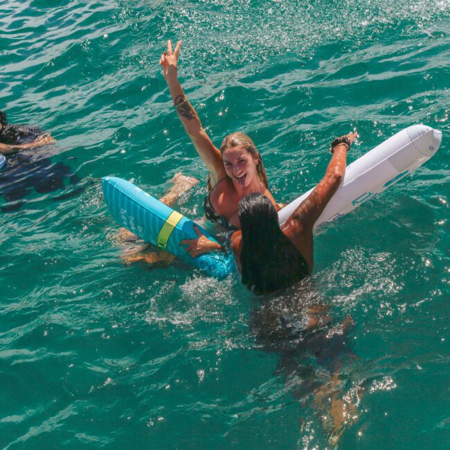Three people are in clear blue water. One woman smiles and flashes a peace sign while holding onto a float, another person holds her, and a man nearby floats with a tray of drinks. "Yacht Social Club" logo is in the corner.