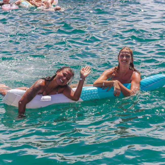Two women smiling and waving while floating on an inflatable raft in clear blue water; other people and floaties are visible in the background, enjoying a sunny day.