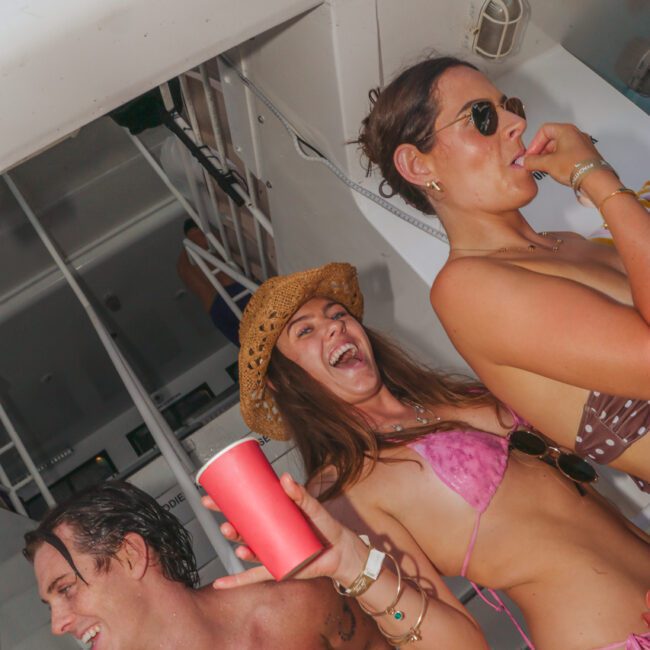 Three people in swimsuits laugh and hold red cups at a lively party on a boat. One wears a straw hat and pink bikini, another sports sunglasses, while the third has wet hair and white face paint. The setting is bright and festive.