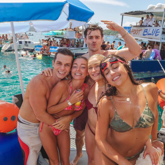 A group of five young adults in swimsuits smile and pose together on a boat in sunny, turquoise water. Other people and boats are visible in the background, and everyone appears to be enjoying a lively day on the water.