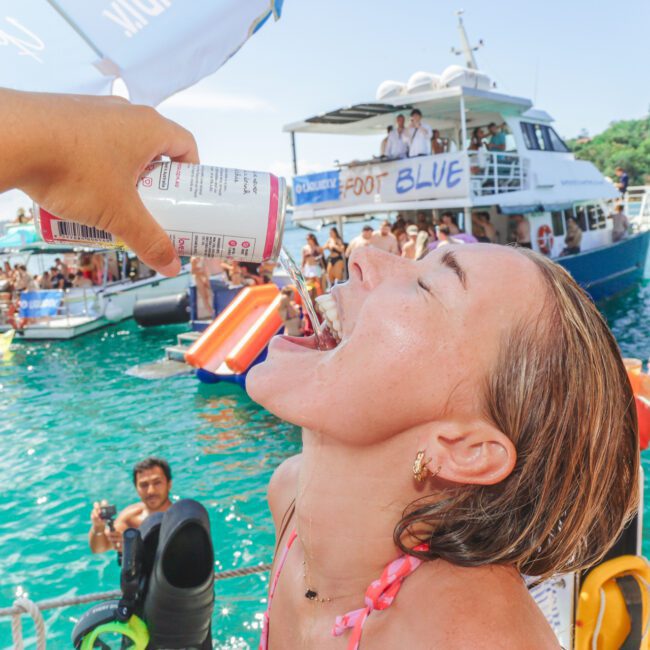 A woman in a bikini smiles and tilts her head back as someone pours a canned drink into her mouth on a boat crowded with people enjoying a sunny day on the water, with another boat and people swimming in the background.