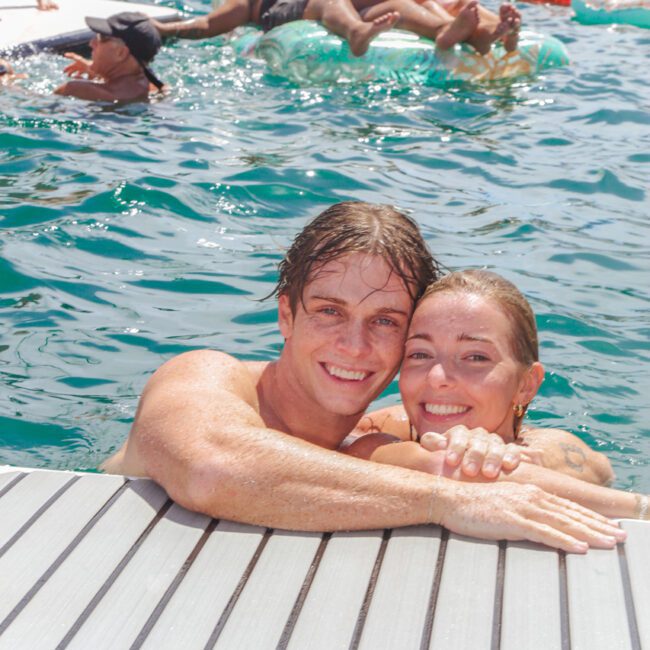 A smiling couple embraces at the edge of a dock in clear blue water, with several people relaxing on inflatables in the background during a sunny day.
