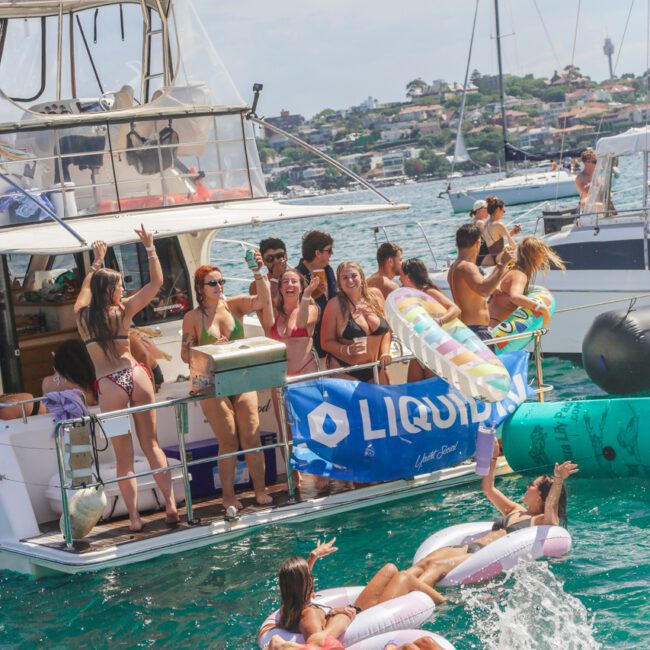 A group of people in swimwear party on a boat, some dancing and cheering, while a few float and splash in the water on inflatable rafts. Other boats and a shoreline are visible in the background.