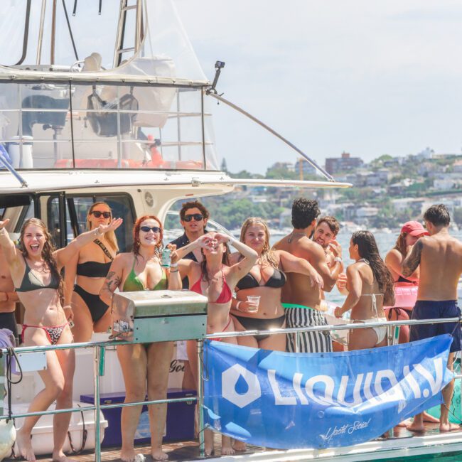A group of people in swimsuits enjoy a party on a boat, smiling, posing, and holding drinks. The boat is docked near the shore with buildings and trees visible in the background on a sunny day.