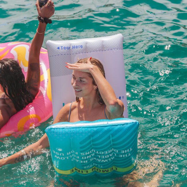 Two women relax on colorful pool floats in clear blue water. One holds the other's hand and looks up while shielding her eyes from the sun. The scene is lively and summery, with a logo in the bottom right corner.