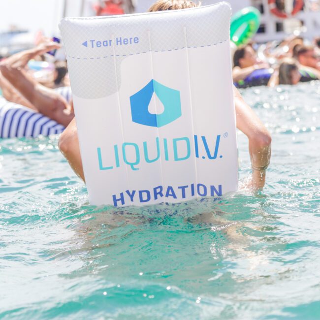 A person in a pool holds a large inflatable mattress with the "Liquid I.V. Hydration" logo, surrounded by others on colorful floaties in the water on a sunny day.