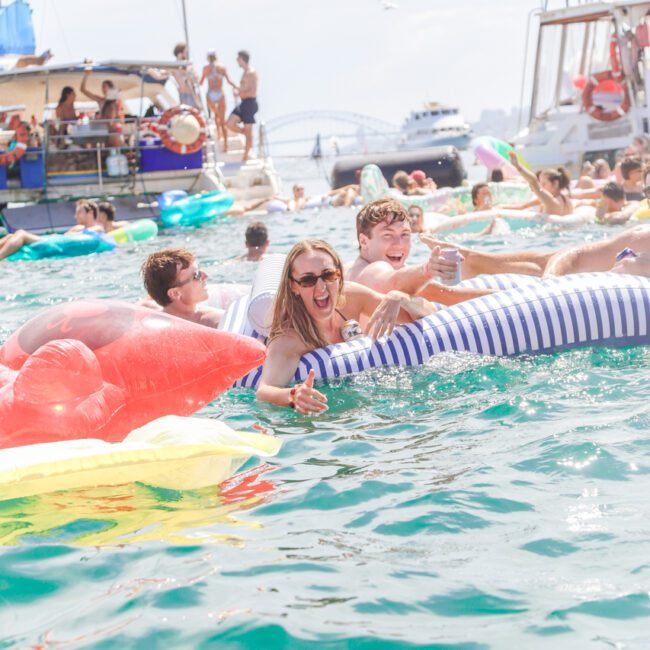 People smiling and relaxing on inflatable floats in the water during a lively boat party, with yachts and more partygoers in the background under a sunny sky.