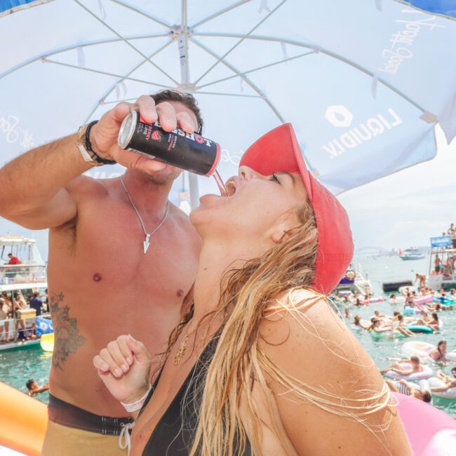 A man pours a drink into a woman's mouth at a crowded lake party. They are under a large umbrella, surrounded by people on inflatables and boats, all enjoying the sunny day.