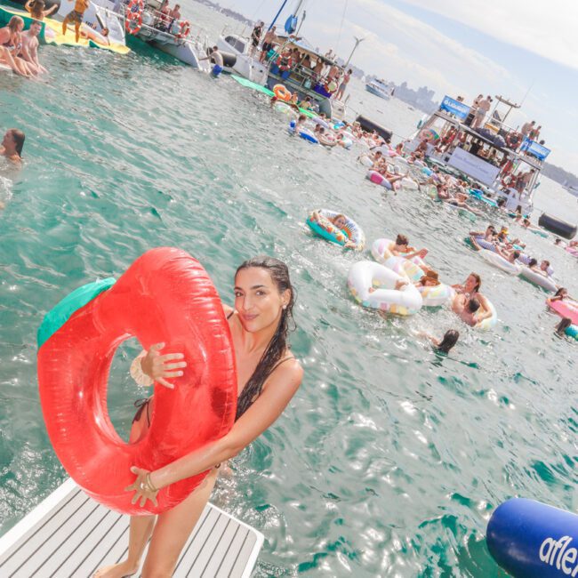 A woman in a swimsuit stands on a boat platform holding a red inflatable ring, smiling at the camera. Behind her, people float on inflatables and gather on boats in a lively, sunny water party scene.