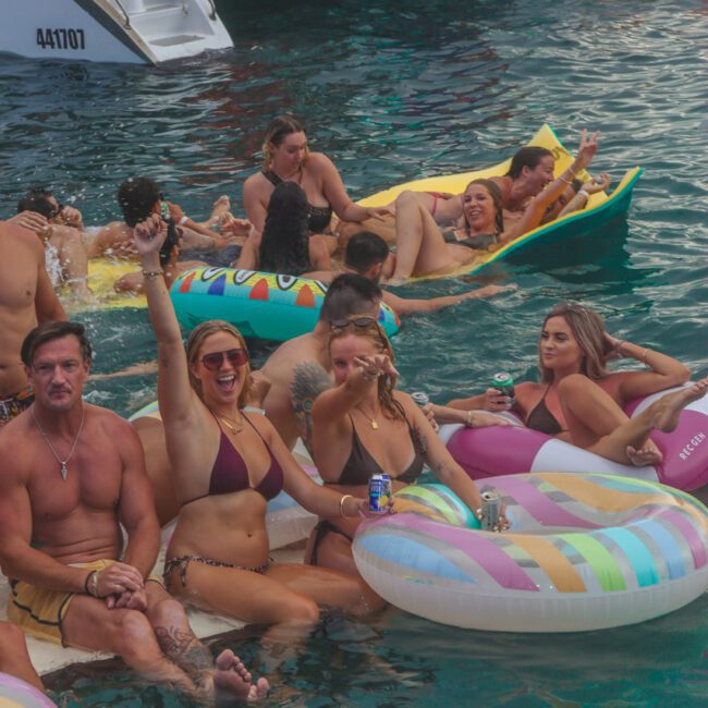 A group of people in swimsuits relax on colorful inflatables and float in the water near a boat, smiling, cheering, and holding drinks at a lively pool party event.