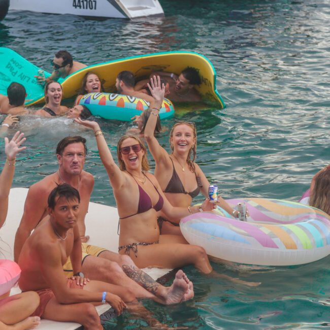 A group of people in swimsuits relax and have fun on colorful inflatables in the water, smiling and waving at the camera. Some hold drinks, and the atmosphere is lively and festive.