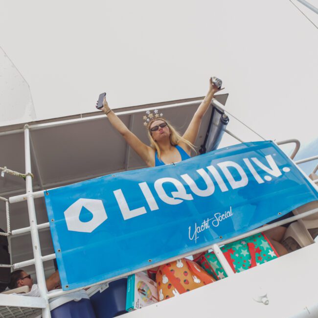A woman wearing sunglasses and a crown stands on a boat deck holding up drinks, with a blue "LIQUID I.V." banner draped over the railing. Colorful gift bags are visible below the banner.