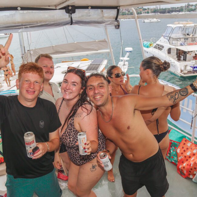 A group of young adults smile and pose with drinks on a boat, wearing swimsuits. Other people relax in the background, with more boats and water visible behind them on a sunny day.