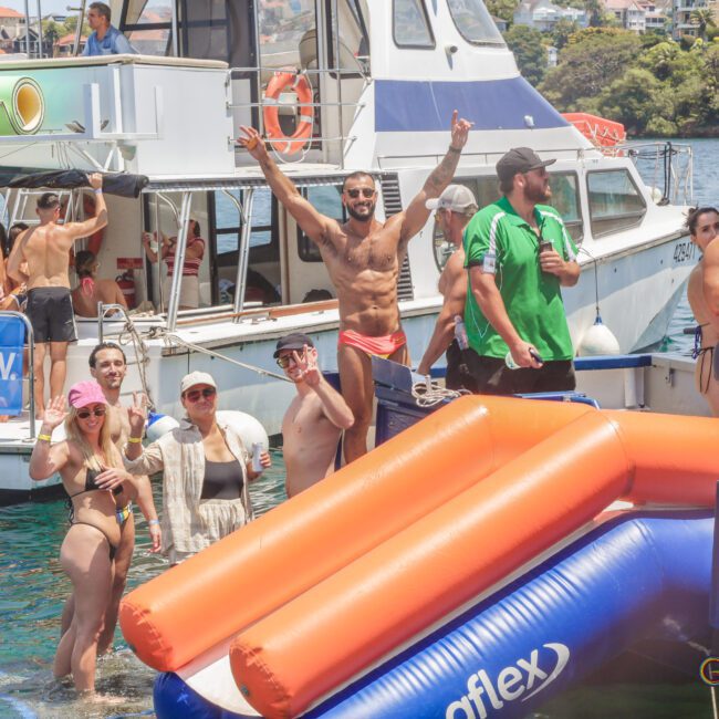 A group of people in swimsuits enjoy a lively boat party on the water, with inflatable floats and slides. Some pose and smile for the camera, while others relax on the boats under sunny weather.