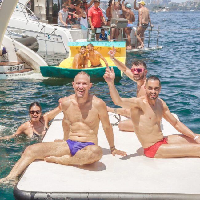 Four smiling people in swimsuits sit and pose on a floating mat in the water, with boats and more people celebrating in the sunny background. The scene is festive and relaxed.