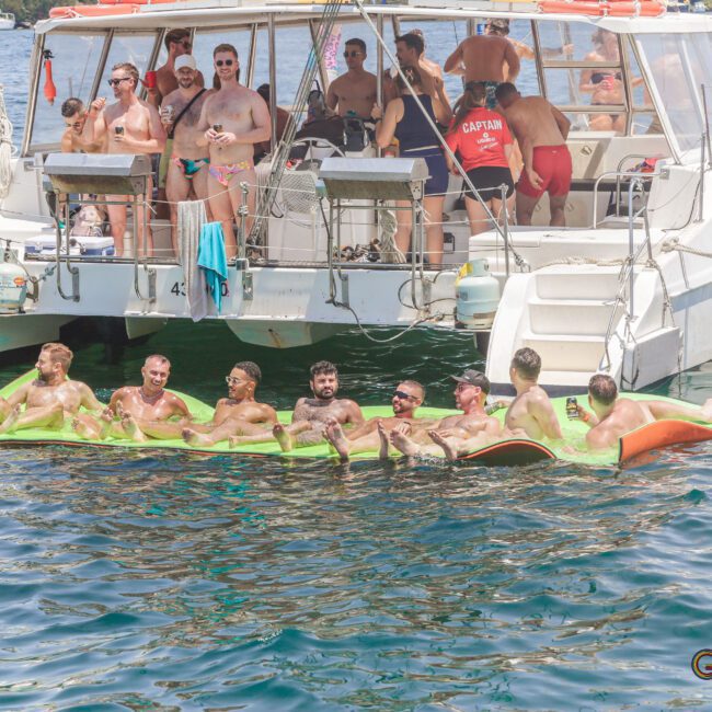 A group of people relax on floating mats in the water near a catamaran, while others stand and socialize on the boat under sunny weather. The scene suggests a fun and lively gathering.