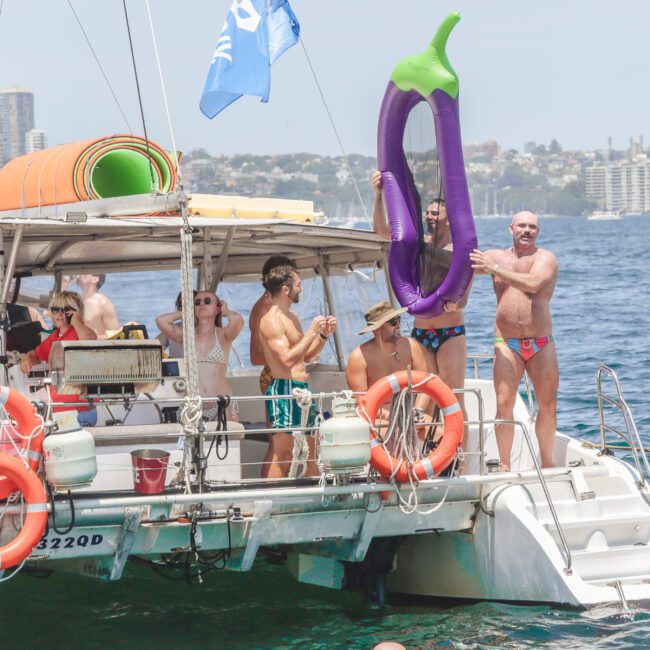 A group of people in swimsuits enjoy a sunny day on a boat; one man stands on the deck holding a large inflatable eggplant. The boat is on the water, with a city skyline and beach visible in the background.