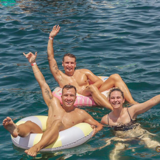 Three people smile and raise their arms while floating in clear blue water on inflatable rings, enjoying a sunny day. The scene looks lively and joyful, with relaxed summer vibes.