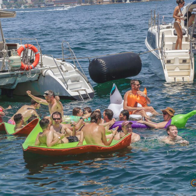 A group of people in swimsuits enjoy a sunny day, floating on colorful inflatables in the sea near anchored boats. Some are laughing, splashing, or talking, and the atmosphere is lively and festive.