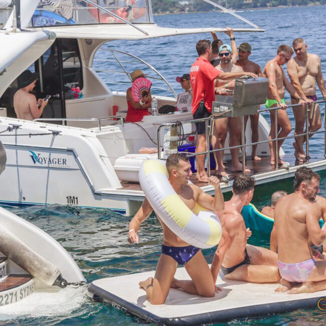 A group of men in swimwear gather on yachts and a floating platform on the water, enjoying a sunny day. One man stands with a swim ring, while others sit or stand, socializing and relaxing by the boats.