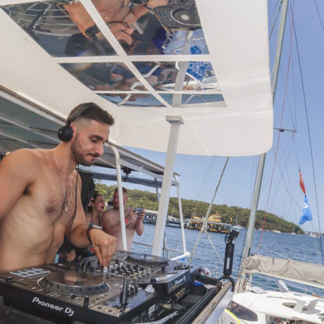 A shirtless DJ wearing headphones mixes music on a Pioneer DJ controller on a boat, with a lively crowd and other boats in the background on a sunny day at sea.