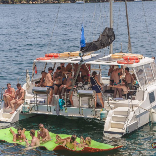 A group of people in swimsuits relax on a white catamaran and on floating mats in the water, enjoying a sunny day. The boat is anchored in calm, clear water near a rocky shoreline.