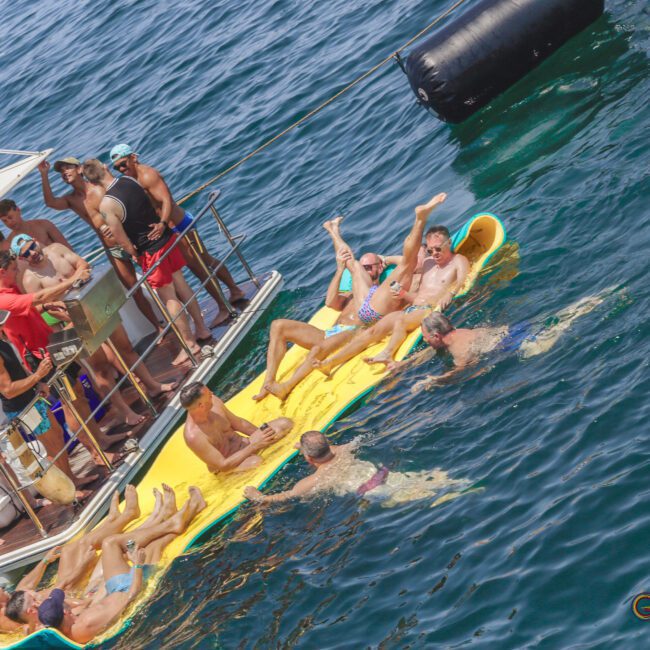 A group of people relax and socialize on bright yellow and green floating mats in the water next to a boat on a sunny day. Some people swim nearby while others enjoy the sun and music.