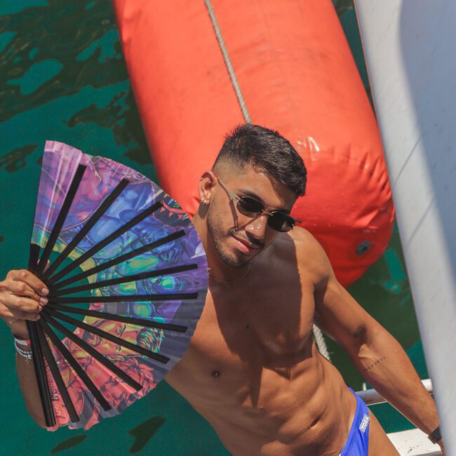 A man in blue swim briefs and sunglasses poses on a boat, holding an open rainbow-colored fan. He is near a large orange buoy floating on green water, enjoying a sunny day.