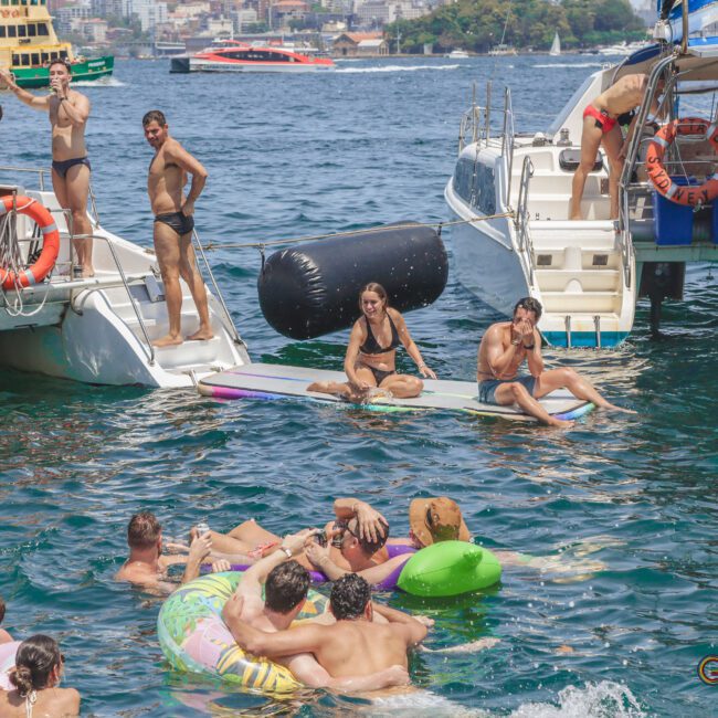 People relax on boats and float on inflatables in a crowded, sunny bay. Some are sitting on a dock between two boats while others swim and socialize in the water, enjoying a lively summer day.